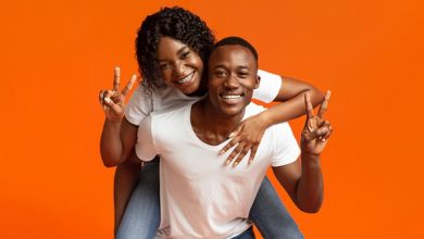 A Black man with short, cropped hair giving a piggyback ride to a Black woman with braces and long wavy hair. They're both in blue jeans and white t-shirts, smiling, and giving a peace sign, the man with his left hand and the woman with her right hand.